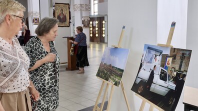 Дві жінки у храмі Різдва Пресвятої Богородиці уважно розглядають фотографії на дерев’яних мольбертах про візит Папи Івана Павла ІІ до Львова. На задньому плані є ікони та інші відвідувачі.