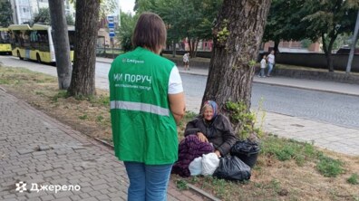 Працівниця соціального патруля спілкується з літньою жінкою, яка сидить біля дерева на узбіччі. Біля жінки лежать речі та пакети, а на задньому плані видно автобус, дорогу й міську забудову. Працівниця одягнена у зелену жилетку з написом «Джерело» та «Ми поруч».