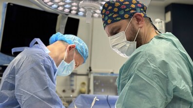 Двоє лікарів у спеціальних медичних халатах, шапочках і масках оперують в операційній, над ними – операційна лампа