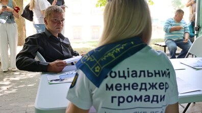 Просто неба за білим столом соціальна менеджерка спілкується з чоловіком. Поруч перебувають інші люди, а на столі лежать інформаційні матеріали та документи. На футболці працівниці видно напис «Соціальний менеджер громади». Фото – із роботи мобільного простору підтримки для потребуючих у Львові.