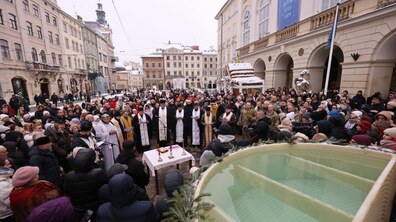 Площа Ринок у Львові. У центрі — велике скупчення людей у зимовому одязі. Посеред площі відбувається традиційне водосвяття: священники у світлих і золотистих ризах моляться біля столу з церковними предметами. Поруч стоять військові. На передньому плані видно велику металеву купіль з освяченою водою, прикрашену зеленими гілками.