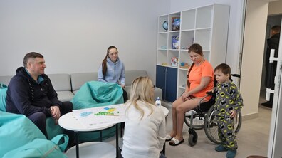 Кілька людей проводять спільне дозвілля у просторій кімнаті тимчасового житла для переселенців на вулиці Замарстинівській. Люди сидять навколо невеликого столу, на якому розкладені різнокольорові деталі гри. Праворуч – жінка у кріслі колісному поруч із маленькою дитиною.
